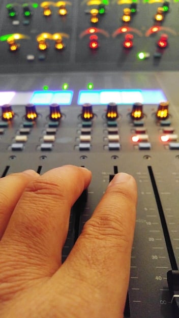 A close-up shot of a sound mixer's hands adjusting the levels on a mixing console, backlit by the glow of the screen displaying film footage. The focus is on the details of the equipment and the precise adjustments being made.