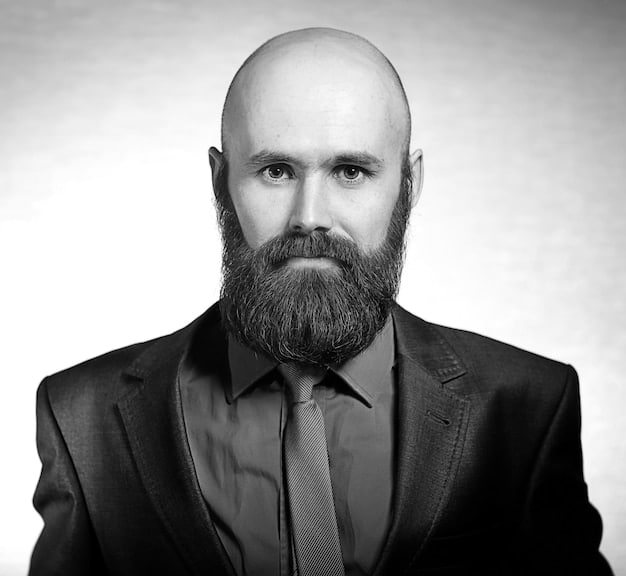 A black and white photograph of Vilhelm Hammershøi, a stern-faced man with a neatly trimmed beard and dark suit, looking directly at the camera, portraying the artist in his younger years.